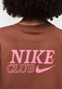 Καφέ βαμβακερό t-shirt με ροζ γραφικό κείμενο που γράφει "NIKE CLUB" και το λογότυπο Nike swoosh στην πλάτη. Κανονική εφαρμογή με στρογγυλή λαιμόκοψη.