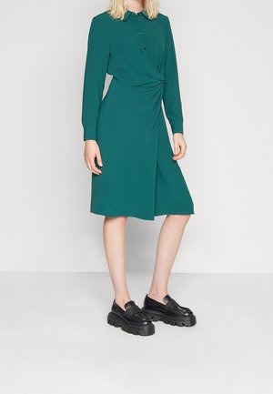 Woman wearing a long-sleeve green knee-length dress with front twist detail and black platform loafers on a plain background.