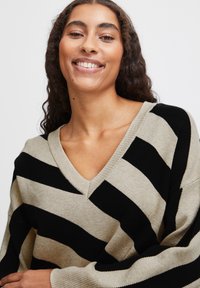 Pull en V avec un motif zigzag beige et noir, fabriqué en matériau tricoté avec une texture douce et une coupe ample.