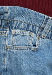 Podrobni posnetek svetlo modrega pasu na jeans hlačah z nabranim elastikom na hrbtni strani in majhno žepom, postavljen proti rdečemu teksturiranemu ozadju.