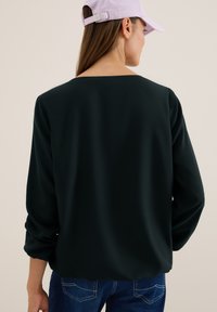 Donkergroene blouse met een wijd decolleté, een losse fit en lange mouwen, gecombineerd met blauwe jeans. Gelaagd textiel en opgerolde mouwdetails.