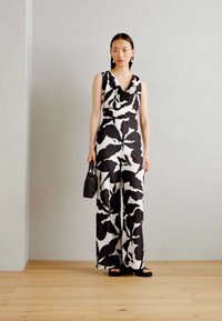 Mono detalle en una camiseta de tirantes de color negro con un escote drapeado que cuenta con un gran patrón de hojas en negro sobre un fondo blanco. La modelo sostiene un pequeño bolso negro.