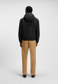 Chaqueta negra con capucha, cierre de cremallera, dobladillo elástico y mangas largas, combinada con pantalones rectos color beige y zapatos negros.