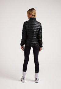 Giacca nera imbottita con collo alto, caratterizzata da un design trapuntato e pannelli laterali elastici, abbinata a leggings neri e scarpe sportive.