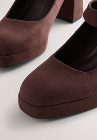 Bruna mockaskor med klossklack, rundad tå, öppna sidor och rem över ankeln. Texturerad yta med sula i slätt material.