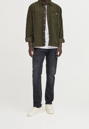 Mies pukeutunut tummanvihreään napilliseen paitaan valkoisen t-paidan päällä, mustiin farkkuihin ja valkoisiin lenkkareihin, seisoo yksinkertaisen vaalean taustan edessä.