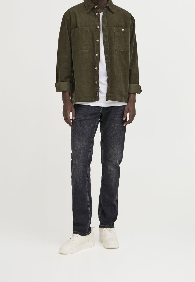 Homme portant une chemise boutonnée vert foncé sur un t-shirt blanc, un jean noir et des baskets blanches, debout devant un fond clair uni.