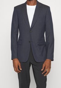 Granatowa, teksturowana marynarka z dwiema przednimi kieszeniami, zapięciem na jeden guzik i klapami w kształcie litery V. Nosi się ją na białej koszulce.