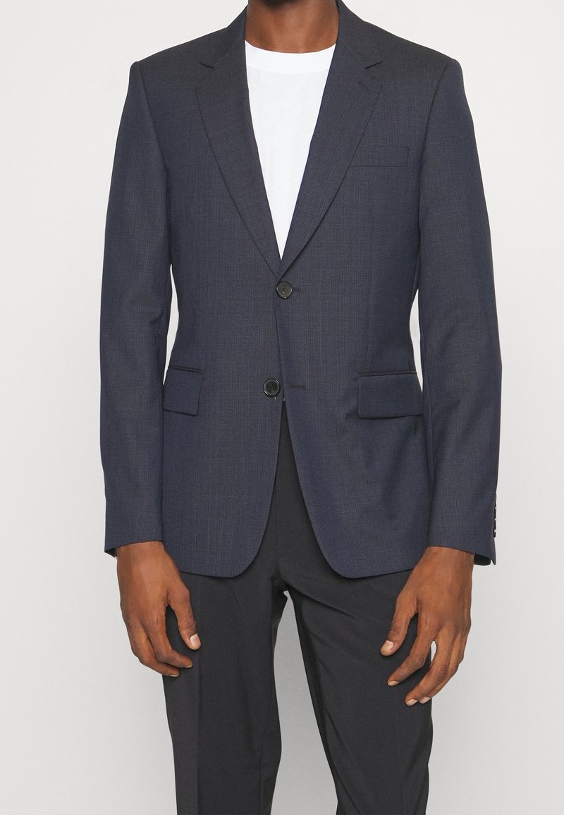 Granatowa, teksturowana marynarka z dwiema przednimi kieszeniami, zapięciem na jeden guzik i klapami w kształcie litery V. Nosi się ją na białej koszulce.