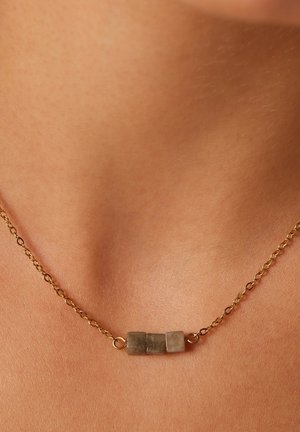 Collier chaîne en or avec trois petites pierres carrées grises centrées sur une peau nue à la base du cou.