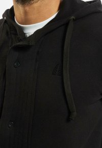 Hoodie nero con una superficie testurizzata, dotato di bottoni, cappuccio con lacci e un sottile logo a foglia. Il tessuto sembra morbido e caldo.