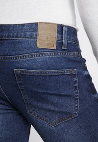 Tumma sininen farkkukangas, jossa on tekstuuri, varustettuna ruskealla nahkalenkillä vyötäröllä, näkyvillä ompeluilla ja takataskulla.