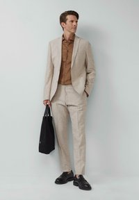 Beige maßgeschneiderter Anzug aus strukturiertem Stoff, kombiniert mit einem braunen Hemd mit Knöpfen. Schwarze Schuhe und eine schwarze Umhängetasche runden das Outfit ab.
