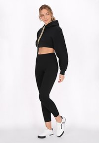Felpa nera a maniche lunghe con cappuccio e dettagli oro, abbinata a leggings neri a vita alta e sneakers bianche con dettagli neri.