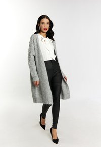 Cardigan lungo grigio con dettagli in perle, indossato sopra una camicetta bianca con fiocco. Abbinato a pantaloni neri attillati e tacchi a punta neri.