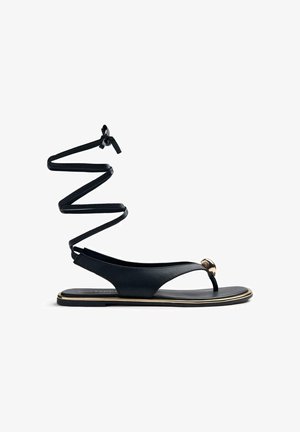 Sandale plate noire avec une bride en T, un accent doré sur la lanière entre les orteils, et de longues brides autour de la cheville nouées en un nœud.