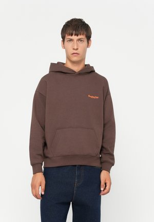 Brown hoodie made of cotton blend. Features a kangaroo pocket, ribbed cuffs, and small orange logo on the chest. Model wears blue jeans.