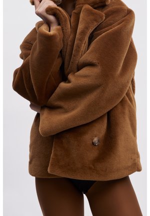 Personne portant un manteau marron en fausse fourrure oversize avec un grand col et un seul bouton visible, mains ajustant le col.