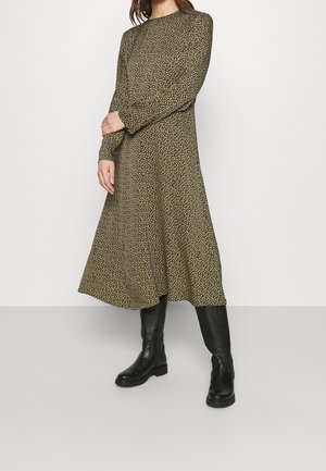 Femme portant une robe midi à manches longues de couleur vert olive avec un petit motif floral, associée à des bottes noires montantes jusqu'aux genoux.