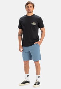 Zwarte katoenen t-shirt met korte mouwen en een groen-oranje graphic op de borst, gecombineerd met lichtblauwe shorts en zwarte sneakers.