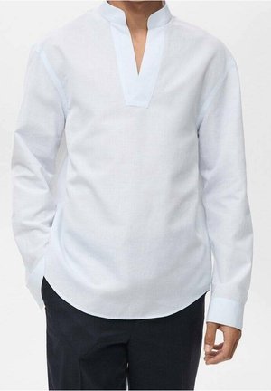 Mand iført en langærmet hvid skjorte med mandarinkrave og sorte bukser, stående med den ene hånd i lommen.