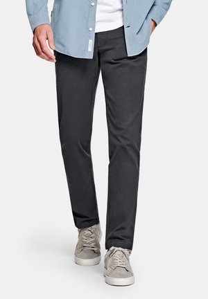 Mann trägt graue Hose, beige Turnschuhe und ein hellblaues Hemd mit Knöpfen, steht vor einem weißen Hintergrund.