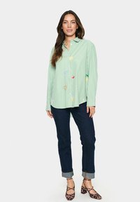 Camicia button-up a righe verdi con ricami di frutta colorata. Indossata con jeans scuri e tacchi con stampa animale. Maniche lunghe, vestibilità comoda.