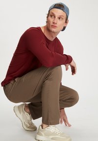 Bordowa koszula z długim rękawem, beżowe slim-fit spodnie i beżowe buty sportowe. Osoba jest w pozycji kucającej, nosi niebieską czapkę z dżinsu. Proste tło.