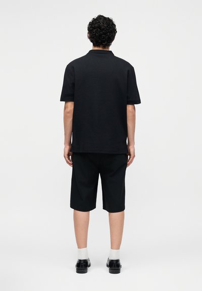 Άνδρας με σγουρά μαλλιά που φοράει μαύρο πόλο με κοντά μανίκια, μαύρο σορτς μέχρι το γόνατο, λευκές κάλτσες και μαύρα παπούτσια, στέκεται με την πλάτη προς τα έξω.