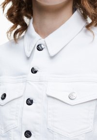 Weiße Jeansjacke mit spitzem Kragen, silbernen Druckknöpfen mit Logo und zwei vorderen Taschen mit Klappen. Strukturierter Stoff.