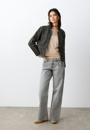 Joven mujer de pie con una blusa beige transparente, chaqueta oscura texturizada, vaqueros gris de pierna ancha, zapatos color ante y un collar rojo ajustado, posando frente a una pared blanca.