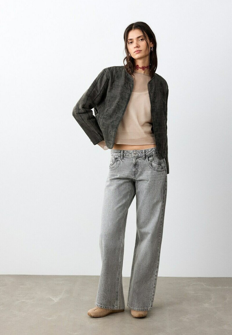 Tânără care stă în picioare, purtând un top bej transparent, o jachetă texturată închisă la culoare, blugi gri largi, pantofi bej și un colier roșu cu choker, în fața unui perete alb.