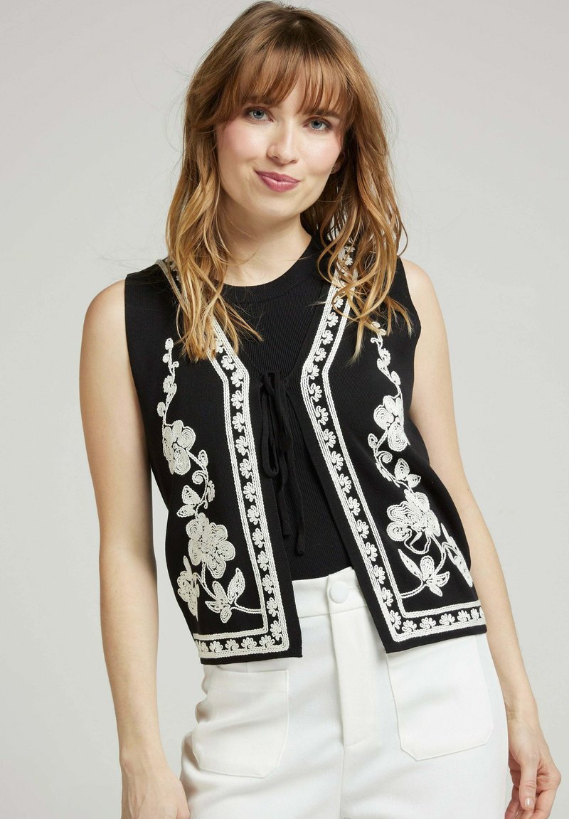 Gilet sans manches noir avec broderie florale blanche, ouvert à l'avant avec un lien, associé à un pantalon blanc taille haute. Texture lisse et design ajusté.