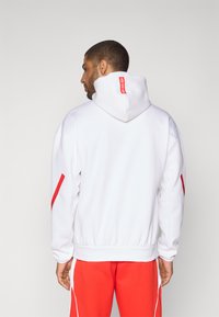 White hoodie with a red accent stripe on the sleeves and a red label on the back of the hood, featuring a ribbed hem and cuffs.