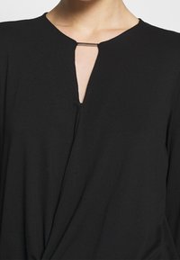 Haut noir avec un décolleté en V et un petit accent argenté au niveau de l'encolure. Fabriqué en tissu léger et texturé avec un design drapé.