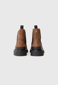 Un par de botas Chelsea de ante marrón con paneles laterales elásticos negros, suelas de goma negra gruesas y lengüetas en la parte posterior.
