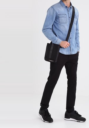 Czarna torba na ramię z pionowymi paskami, wykonana z materiału tekstylnego, prostokątna forma. Nosi ją osoba w jasnoniebieskiej koszuli dżinsowej i czarnych spodniach.