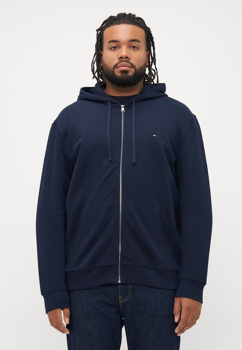 Hoodie zippé marine en tissu doux, avec une poche kangourou, des cordons de serrage et un petit logo sur la poitrine. Poignets et ourlet texturés.