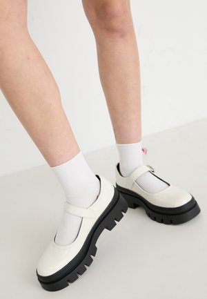 Zapatos Mary Jane de charol blanco con una gruesa plataforma negra y correa. Usados con calcetines blancos hasta el tobillo. Suela negra texturizada con surcos.