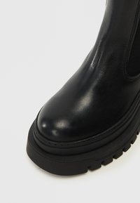 Crne kožne ankle čizme s okruglim vrhom, debelim gumenim potplatom i elastičnim bočnim panelima. Glatka tekstura s prošivenim detaljima na gornjem dijelu.