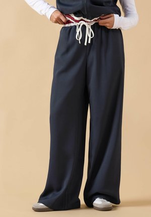 Person trägt locker sitzende dunkelblaue Hose mit weiß-rot gestreiftem elastischem Bund und weißem Kordelzug, kombiniert mit weißen Schuhen.