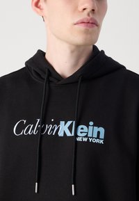 Black hoodie made of cotton blend, featuring a drawstring hood, front logo in white and light blue, and silver hardware on drawstrings.