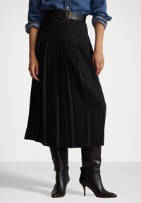 Gonna nera plissettata con vita alta e design con cintura, abbinata a stivali con tacco neri lucidi. Il tessuto appare liscio e sartoriale.