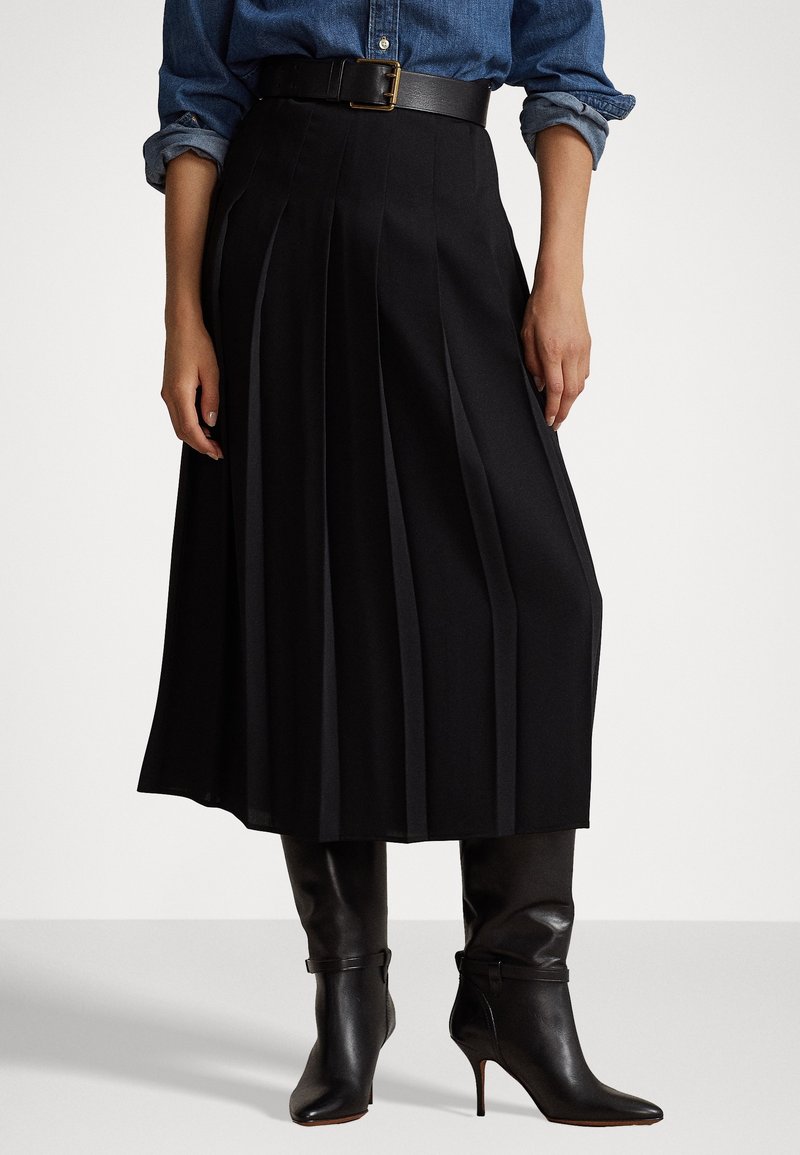 Gonna nera plissettata con vita alta e design con cintura, abbinata a stivali con tacco neri lucidi. Il tessuto appare liscio e sartoriale.