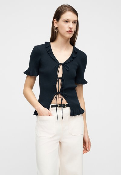 Νεαρή γυναίκα με ίσια καστανά μαλλιά φοράει μαύρο τοπ με κοντά μανίκια, δεσίματα μπροστά και βολάν στις άκρες, συνδυασμένο με λευκό παντελόνι ψηλής μέσης.