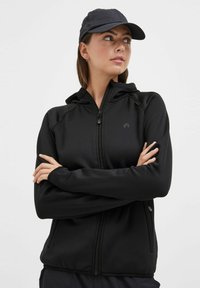 Zwarte rits-hoodie met een slim-fit design, voorzien van een capuchon met trekkoord en een zachte textuur. Gecombineerd met een donkere pet voor extra functionaliteit.