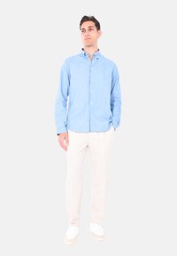 Camicia azzurra chiaro con maniche lunghe, abbinata a pantaloni beige e sneakers bianche. La camicia presenta un colletto classico e un orlo curvo.