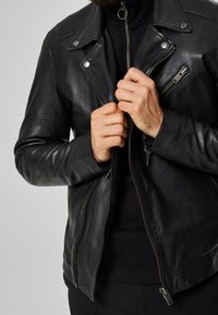 Man som har på sig en svart läderjacka och svart skjorta, som drar upp dragkedjan på jackan med båda händerna.