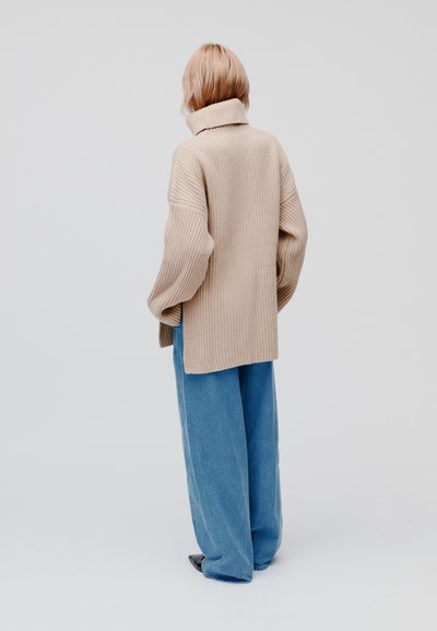 Beige geribbelde trui met een hoge kraag, oversized pasvorm en lange mouwen, gecombineerd met lichtblauwe wijde jeans en zwarte schoenen.