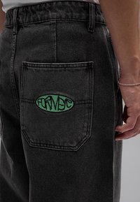 Vaqueros negros de mezclilla con un parche ovalado verde y texto estilizado negro en el bolsillo trasero, usados por una persona con una pulsera plateada.